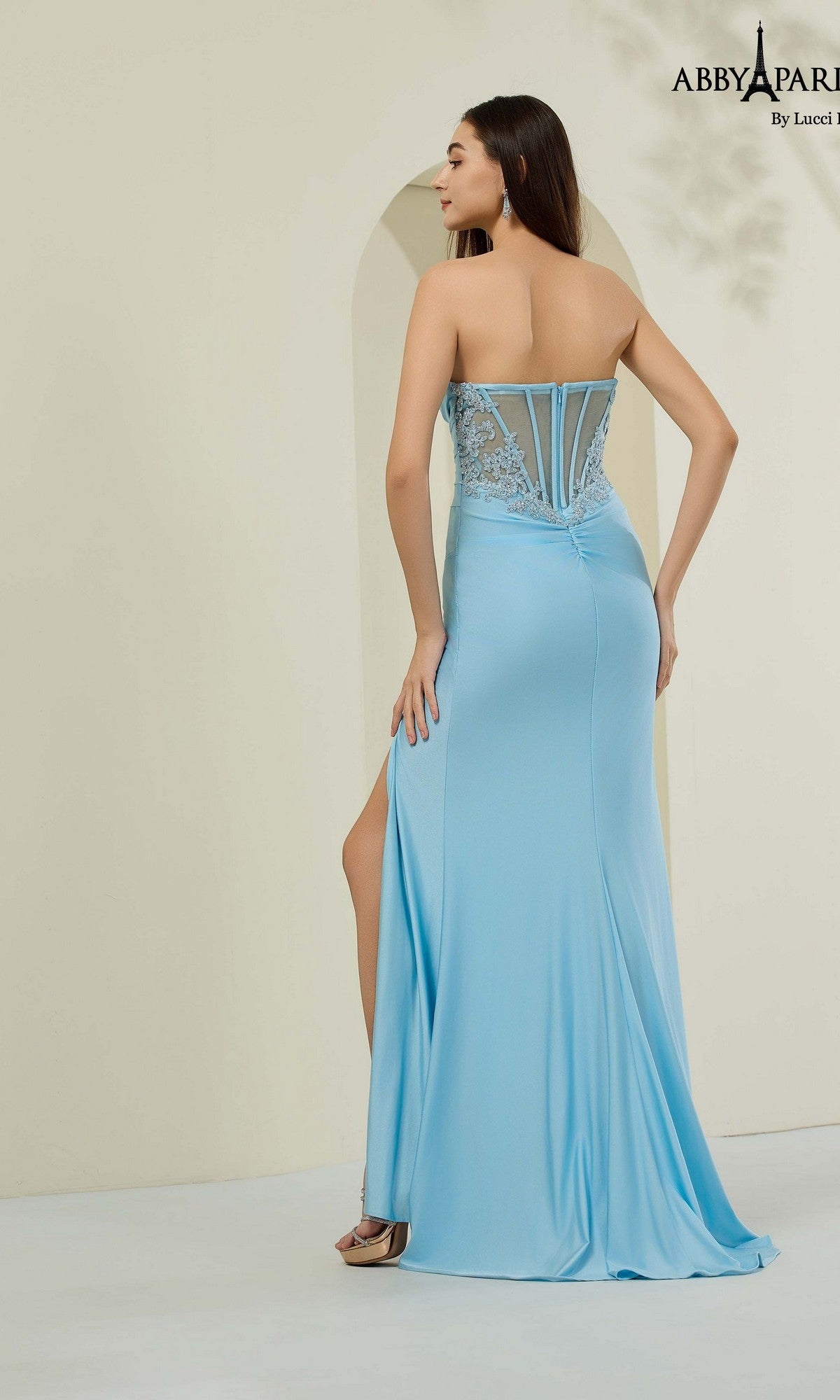 A woman stands with her back to the camera wearing the Abby Paris 90361 long prom dress, featuring a strapless design in light blue with lace detailing and a high slit.